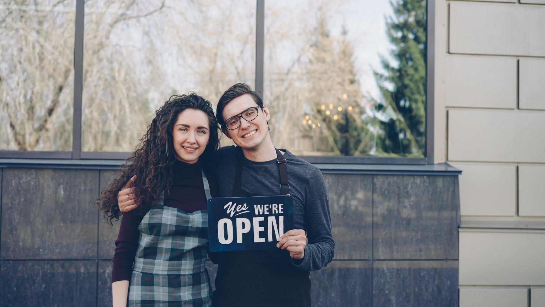 Smiling small-business owners standing outside their shop with an open sign